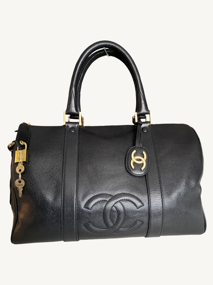 A black leather Chanel duffle bag with gold-tone hardware, featuring the embossed Chanel logo on the front, a top handle, and a lock with keys hanging from the side.