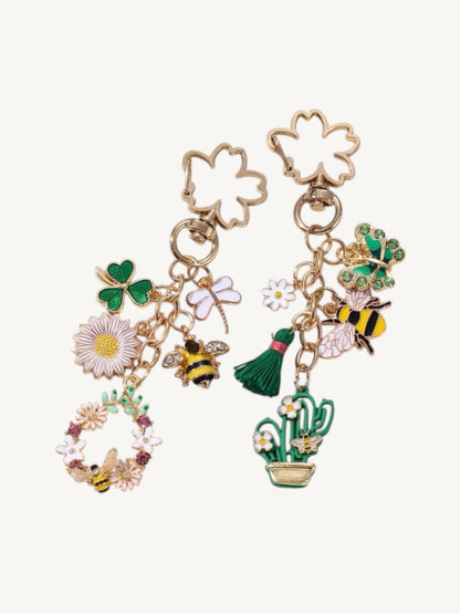 A pair of gold-tone dangling earrings with St. Patrick's Day themed charms, including shamrocks, flowers, bumblebees, a broom, and a cactus, shown on a light cream background.
