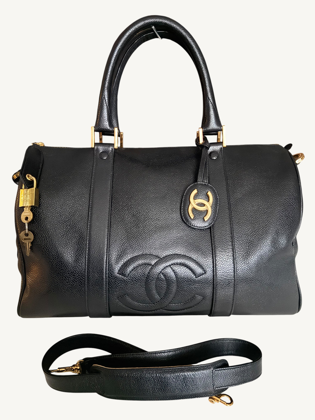 A black pebbled leather Chanel duffle bag with gold-tone hardware, featuring the embossed Chanel logo on the front, a top handle, a detachable shoulder strap, and a small padlock with key hanging from the side, shown against a light off-white background.