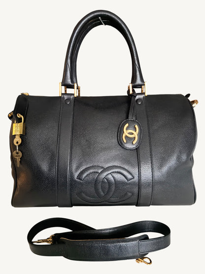 A black pebbled leather Chanel duffle bag with gold-tone hardware, featuring the embossed Chanel logo on the front, a top handle, a detachable shoulder strap, and a small padlock with key hanging from the side, shown against a light off-white background.