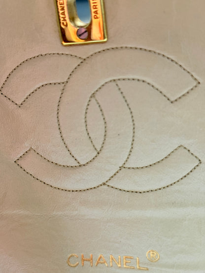 Close-up of a light beige Chanel leather detail featuring a stitched double-C logo, a gold-tone metal tag with 'CHANEL PARIS' text, and the gold 'CHANEL' wordmark at the bottom.