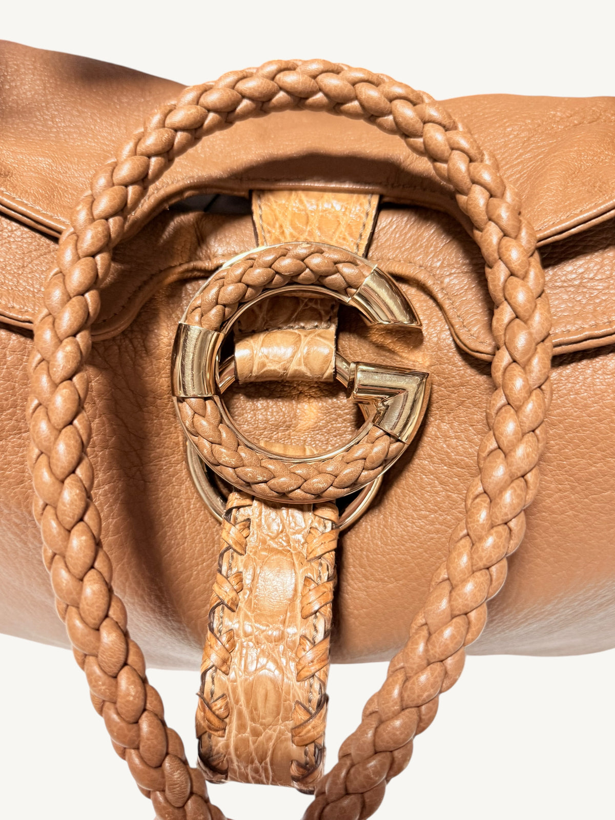 Close-up of a camel brown Gucci  leather handbag featuring a braided shoulder strap, a matching braided strap detail, and a large interlocking G-shaped metal clasp on a light background.