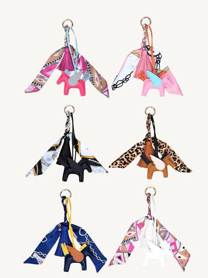 Six fashion horse-shaped keychains arranged in a 3x2 grid on a light cream background. Each keychain features a small horse charm in pink, black, brown, blue, or white, attached to a matching scarf wing with a gold-tone key ring.