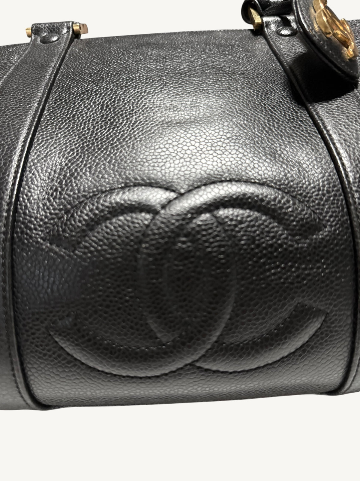 Close-up of a black textured leather handbag with an embossed interlocking CC logo, showing the Chanel brand identity and gold-tone hardware against a light background.