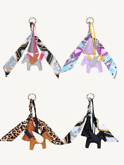 Four fashion keychains featuring small horse charms and patterned scarf wings, arranged in a 2x2 grid on a light cream background.