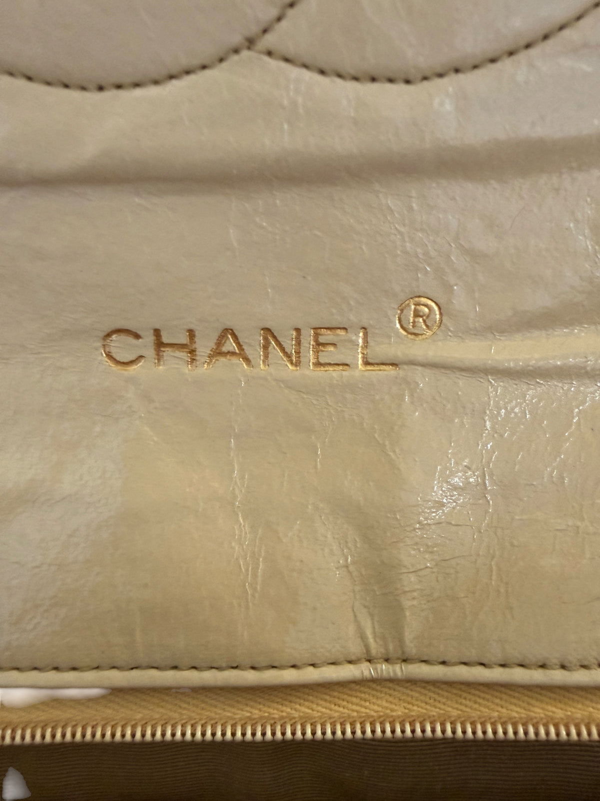 Close-up of a beige Chanel showing the gold-embossed CHANEL logo on textured leather, with a gold zipper along the bottom.