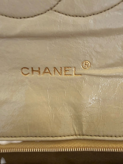 Close-up of a beige Chanel showing the gold-embossed CHANEL logo on textured leather, with a gold zipper along the bottom.