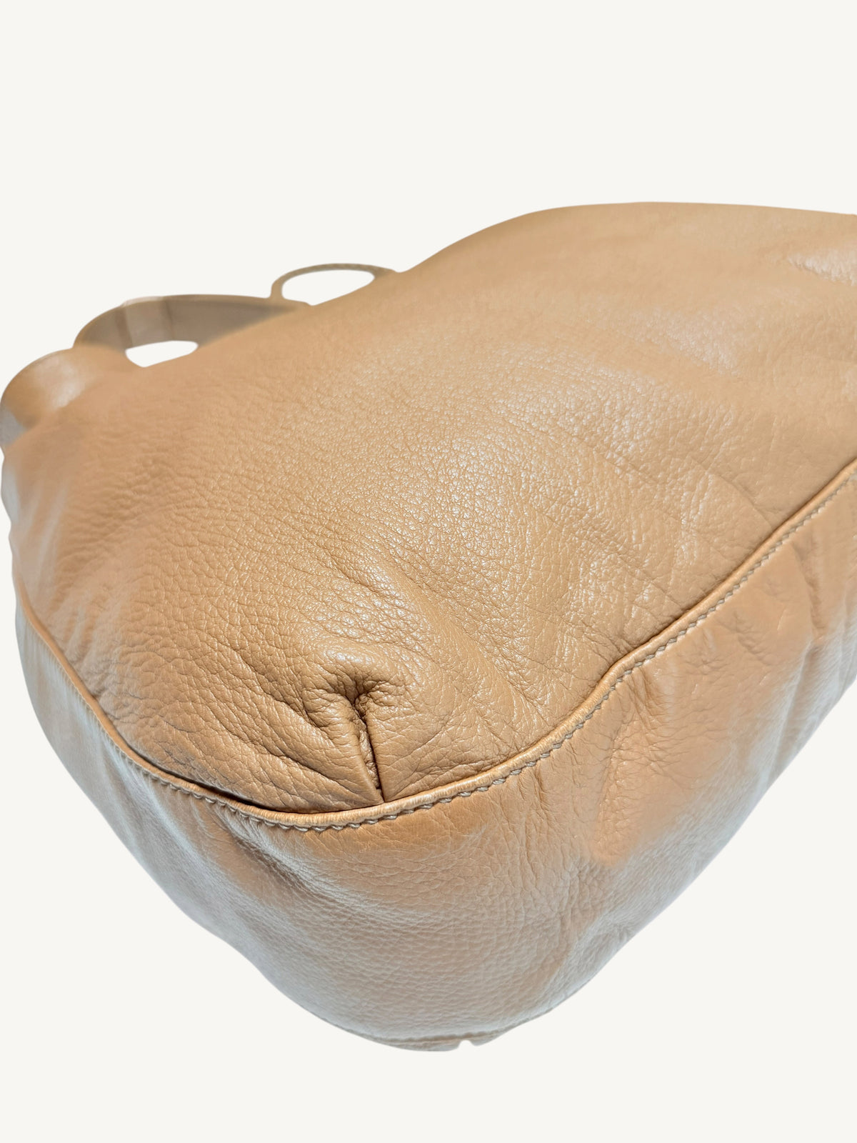 A close-up of a camel brown Gucci  leather shoulder bag showing the textured grain and stitching on a plain off-white background.