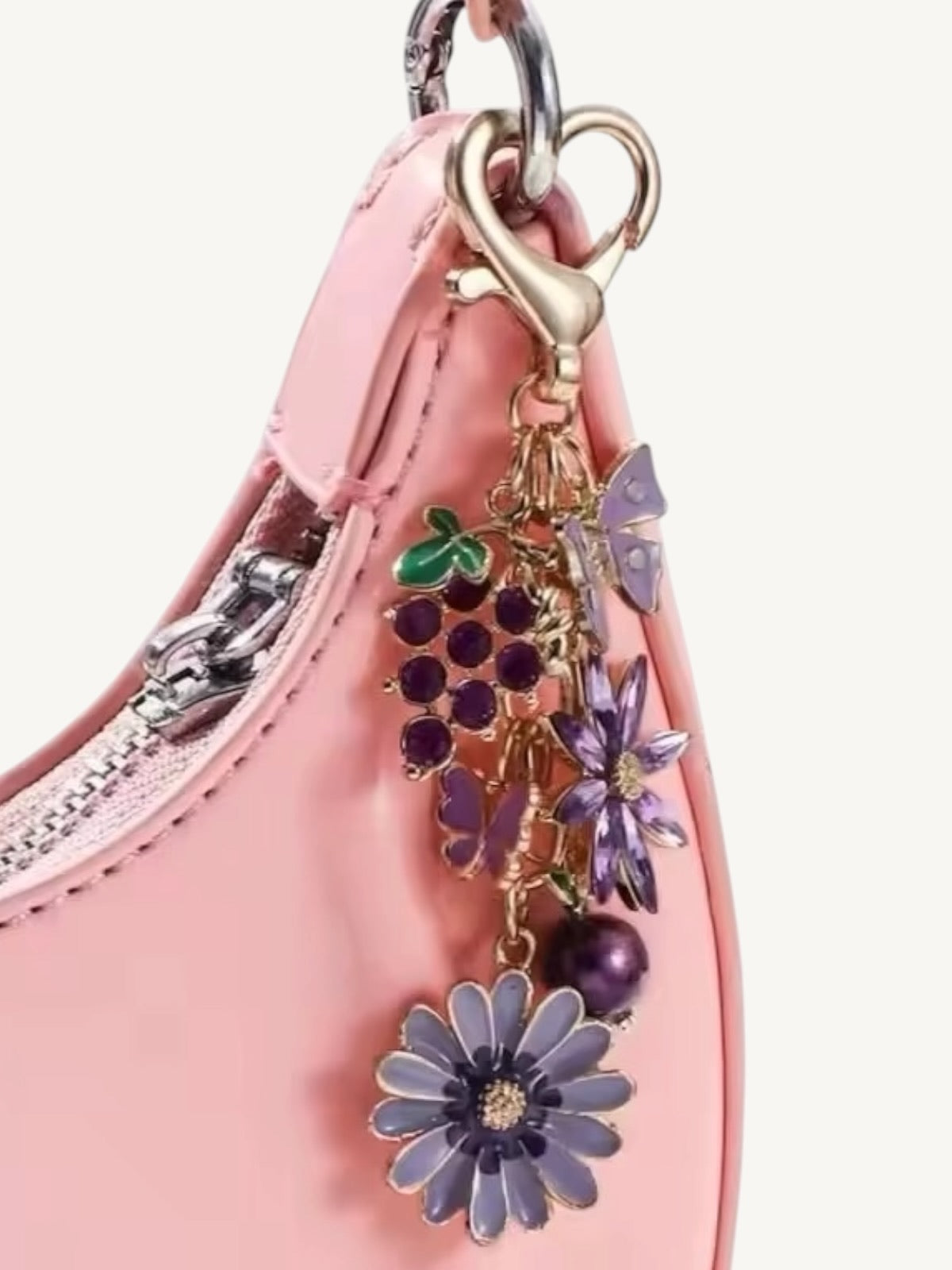 A close-up of a light pink leather handbag charm featuring purple flower charms, purple grape clusters, a butterfly, and a heart-shaped gold clasp.