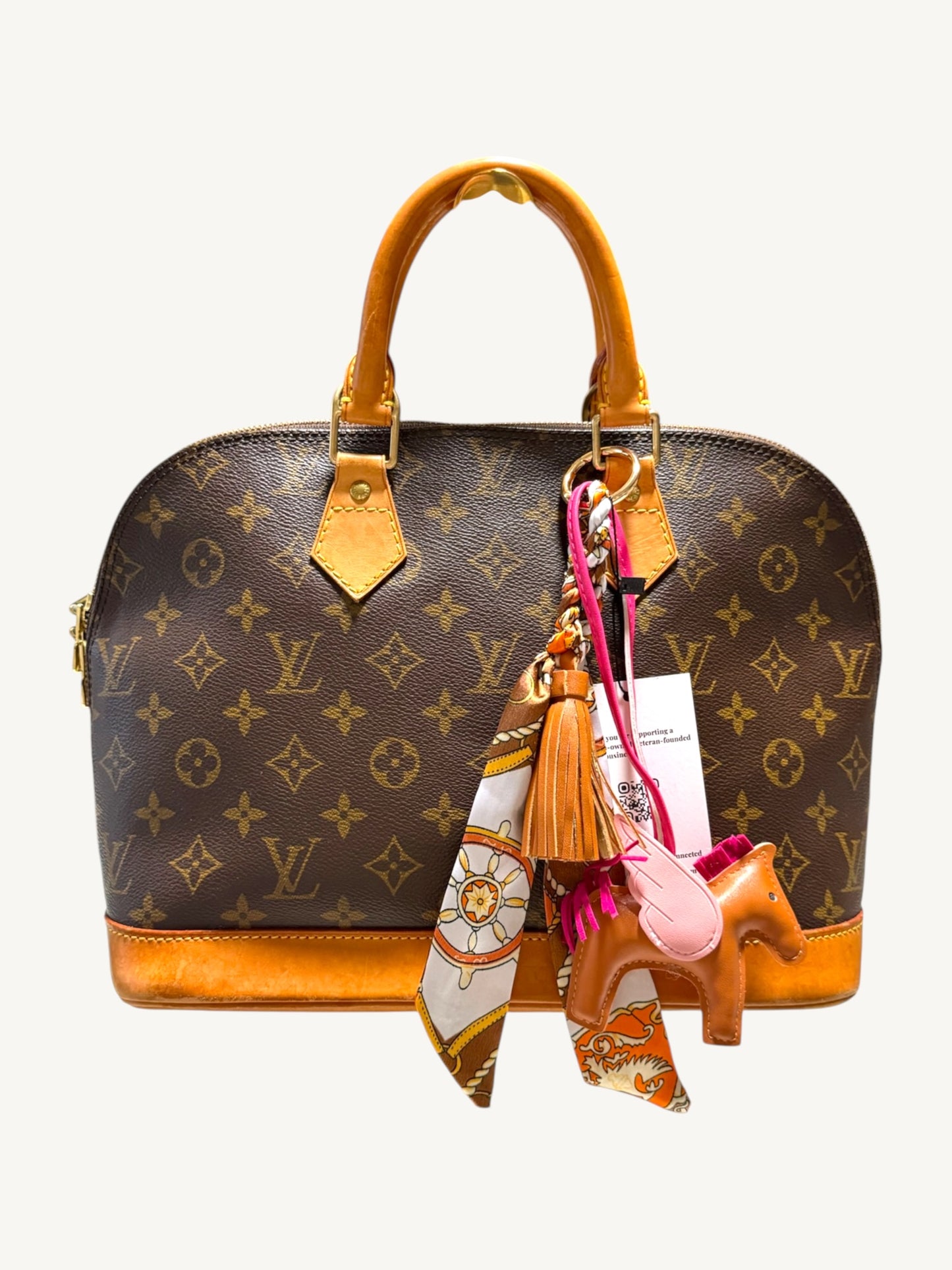 Louis Vuitton monogram canvas dome handbag with tan leather handles and base, adorned with a pink and orange patterned scarf, tassel, and a small horse-shaped charm, shown against a plain light background.