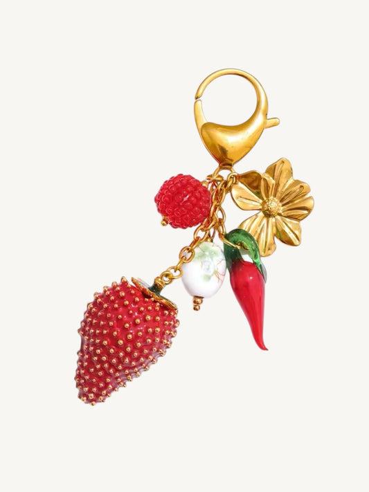 A gold-tone charm keychain featuring a textured red strawberry, a red raspberry, a red chili pepper with green leaves, a white pearl bead, and a gold-tone flower, all attached to a gold lobster clasp on a light off-white background.