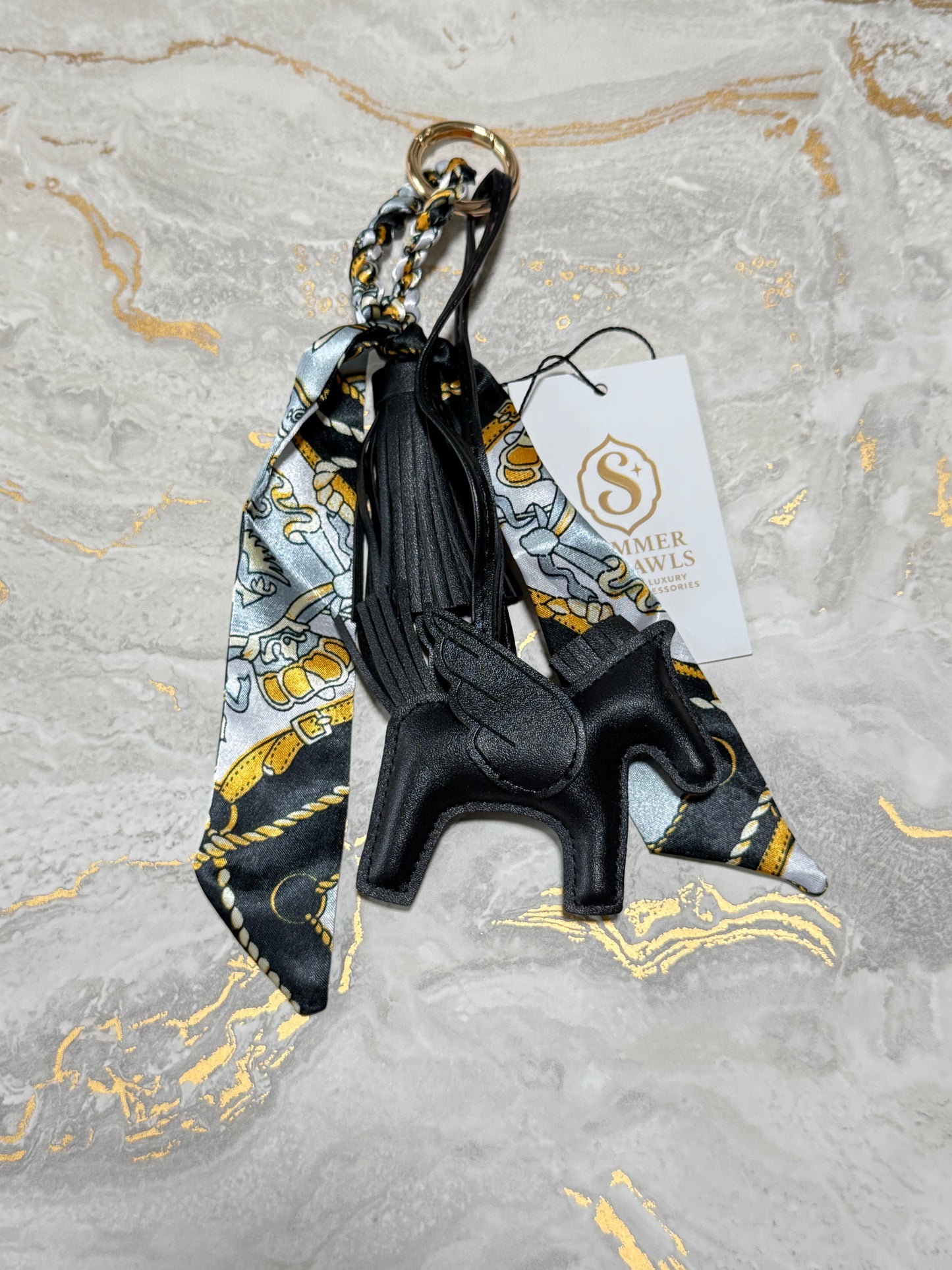 A black leather winged horse keychain charm with a matching patterned silk scarf and a white brand tag, displayed on a gray marble surface with gold veins.