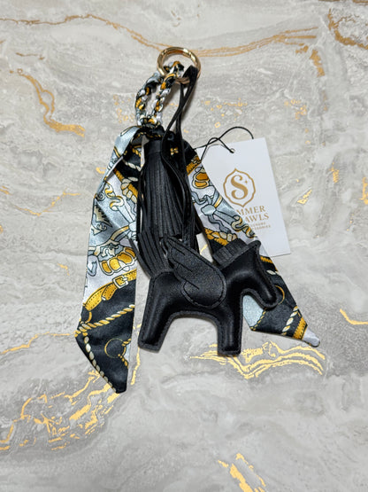 A black leather winged horse keychain charm with a matching patterned silk scarf and a white brand tag, displayed on a gray marble surface with gold veins.