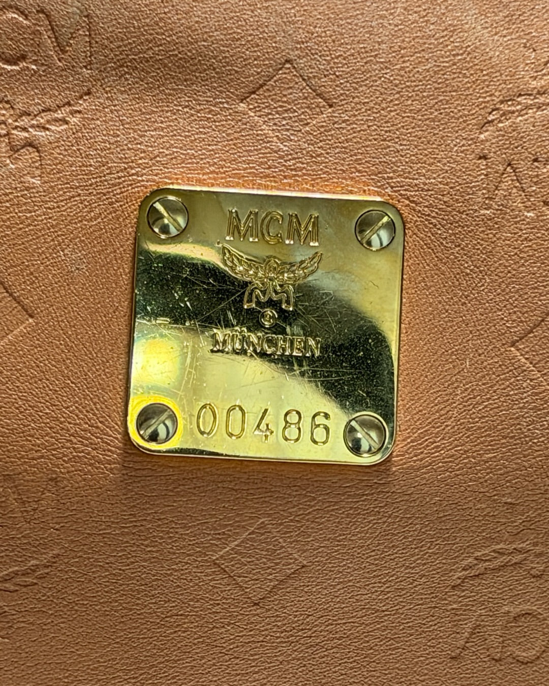 Close-up of scratched gold-tone MCM München plaque numbered 00486 on cognac leather.